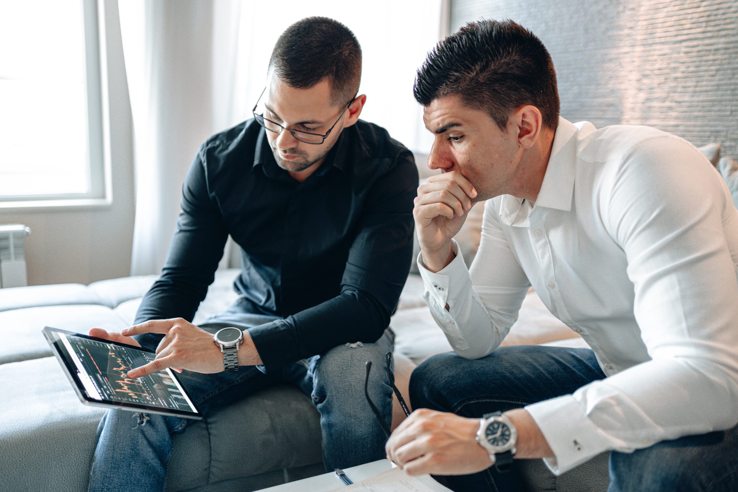 two business men sitting side by side looking at a tablet; one holding it and pointing to a graph it displays while the other leans over to see the screen