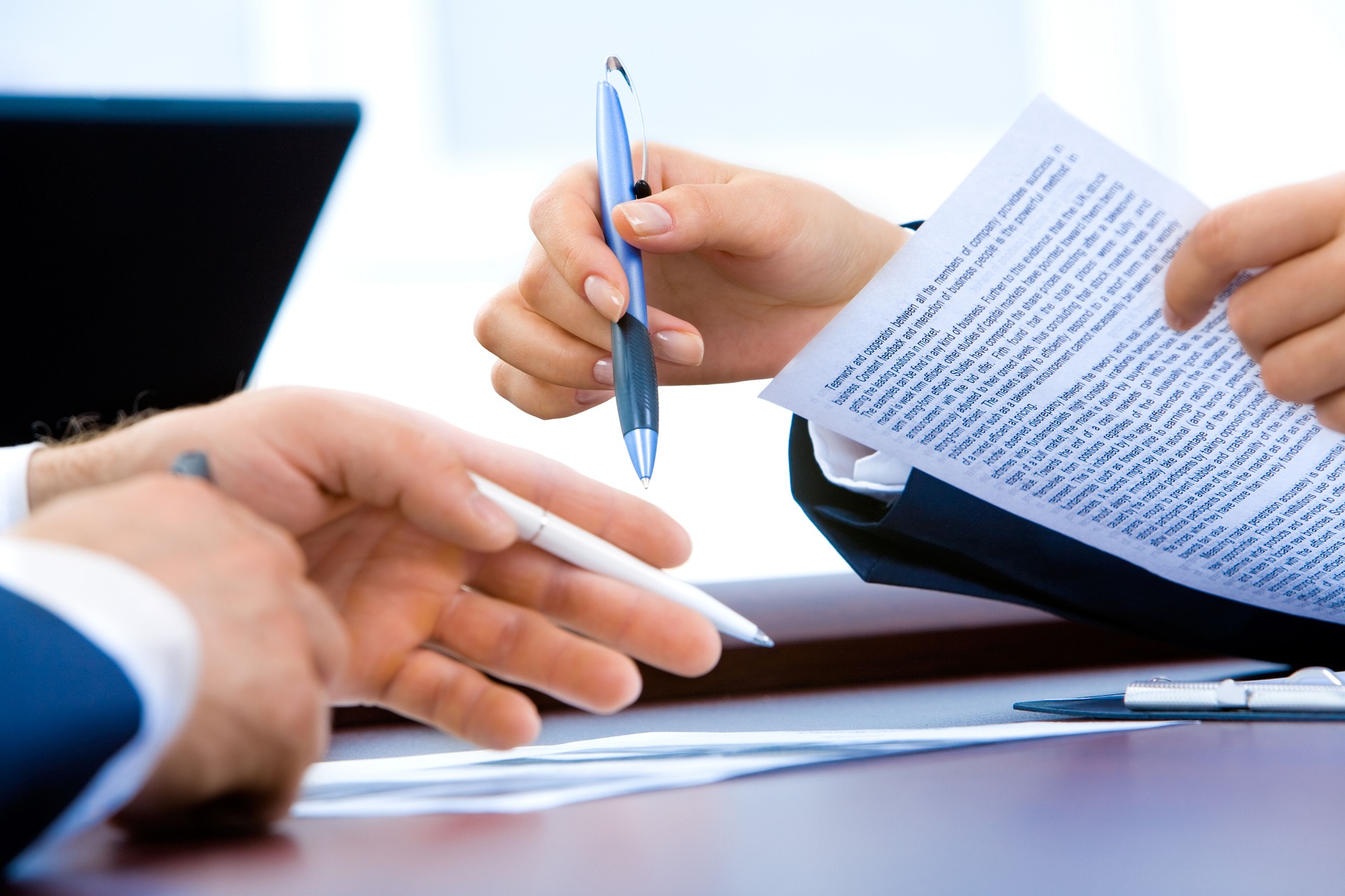 two businesspeople opposite each other at a desk looking over documents with pens in hand and a laptop nearby